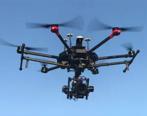 A black drone with six propellers soars against a clear blue sky, perfect for capturing stunning aerial photography. Equipped with a mounted camera and featuring sleek red accents, it promises remarkable views and vivid imaging from above.