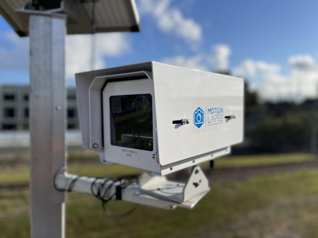 A large white security camera mounted on a pole, labeled with the MOTION LAPSE logo, oversees the scene. Its designed for time-lapse surveillance for construction, set against a blurred background of grass and a building under a clear blue sky with clouds.