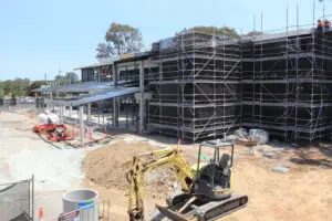 Construction site with a partially built two-story building wrapped in scaffolding. Various construction materials and equipment are scattered around. A small excavator is in the foreground, and trees are visible in the background.