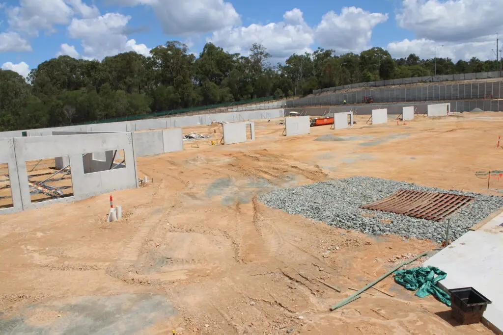 A construction site with a sandy ground and scattered concrete wall panels. Gravel and a metal grate are visible in the foreground. Trees line the background under a blue sky with white clouds.