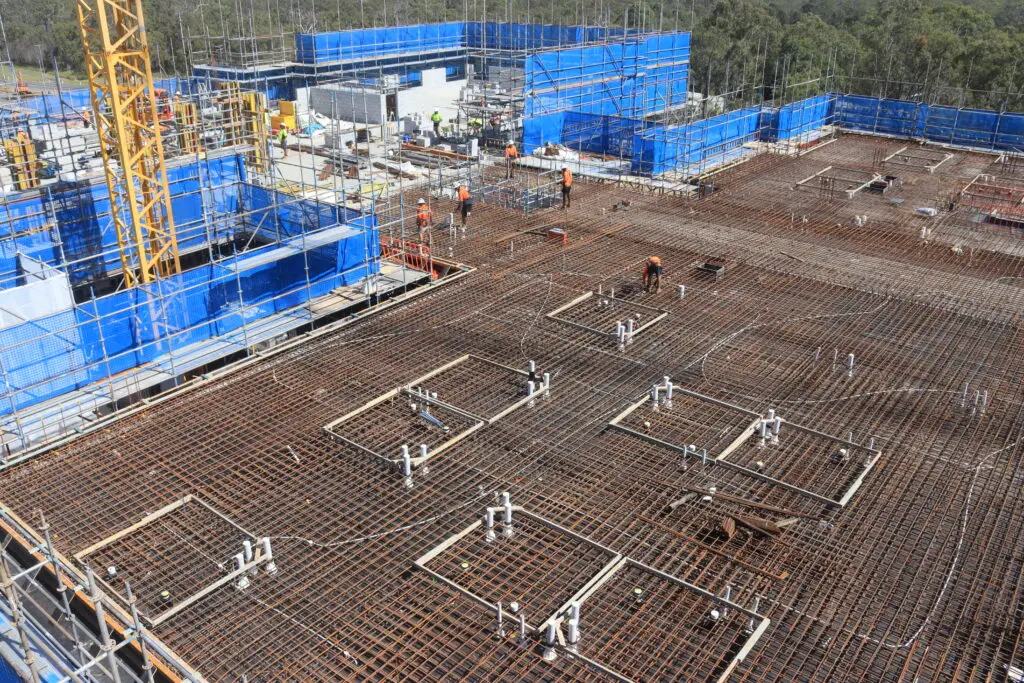 Construction site with a wide view of a building foundation. Steel reinforcement bars are visible, with several workers in orange vests and hard hats. Blue safety barriers and a yellow crane are in the background, surrounded by trees.