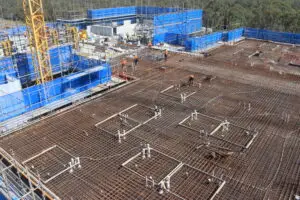 Construction site with a wide view of a building foundation. Steel reinforcement bars are visible, with several workers in orange vests and hard hats. Blue safety barriers and a yellow crane are in the background, surrounded by trees.
