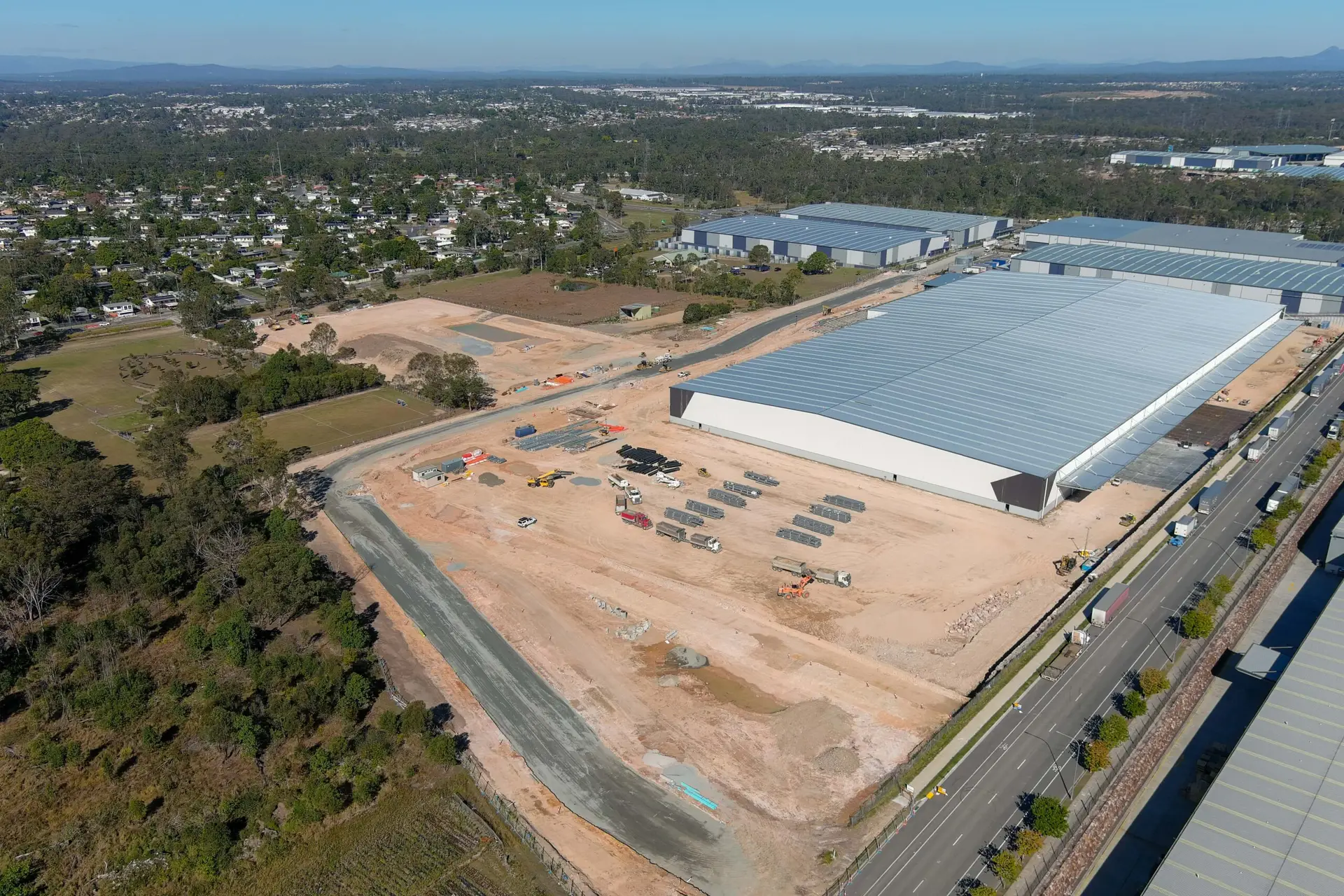 An aerial view of a large construction site adjacent to industrial buildings. The site is partially cleared with construction equipment and scattered materials. Roads run alongside, and trees and a residential area are visible in the background.