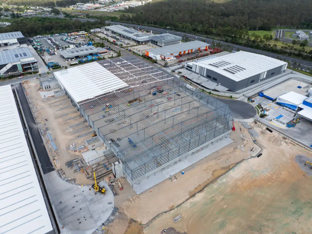 Aerial view of an industrial area under construction. A large warehouse structure with a partially built metal roof dominates the scene. Surrounding buildings and parking lots are visible, with trees in the background. Construction equipment is scattered around.